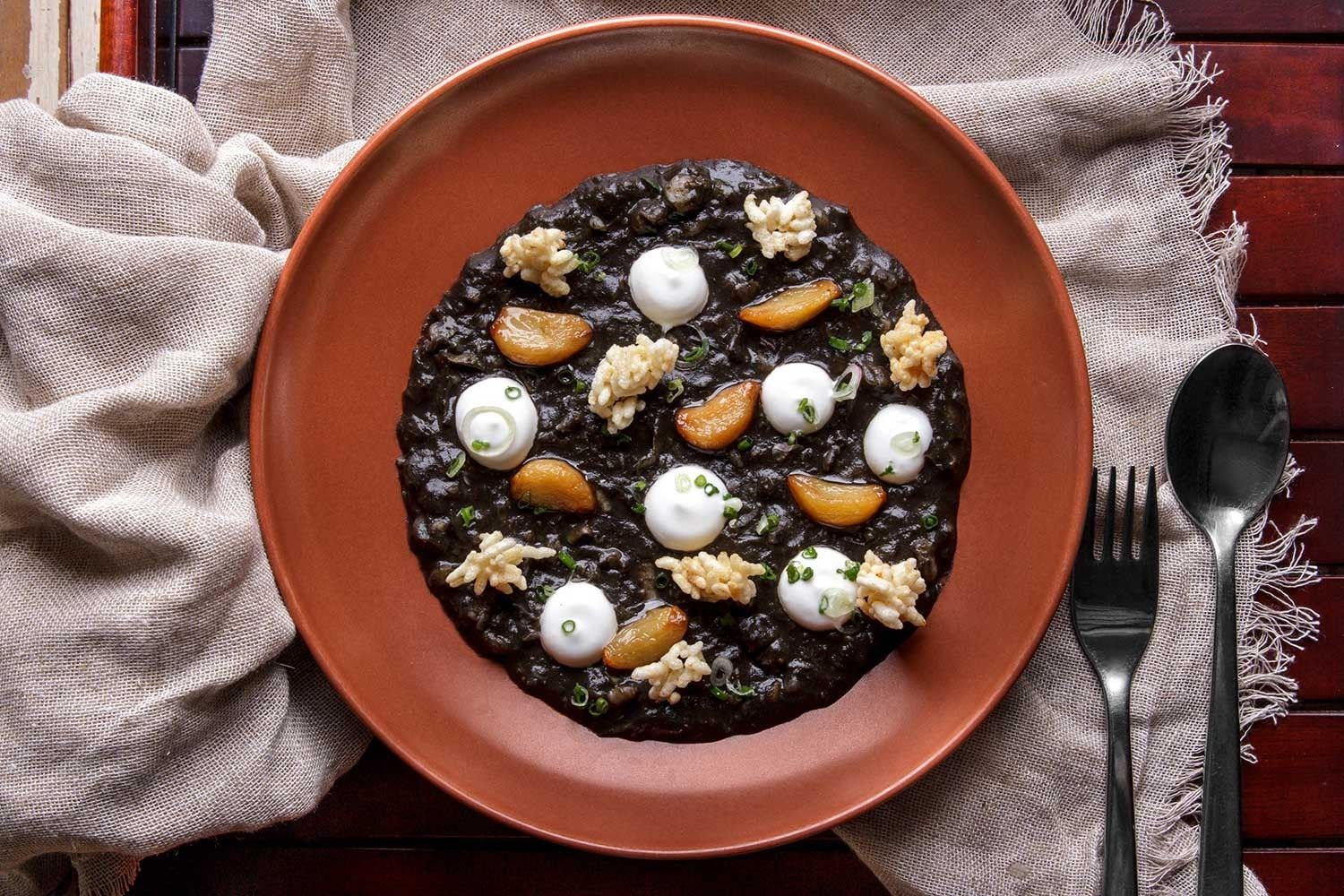 A dark risotto-style dish with small garnishes served on a terracotta plate at Cuca
